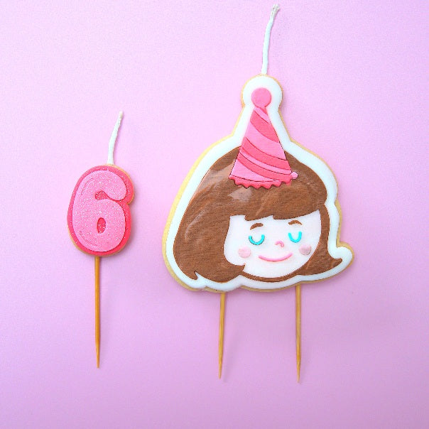A cookie candle of a girl with brown hair wearing a party hat
