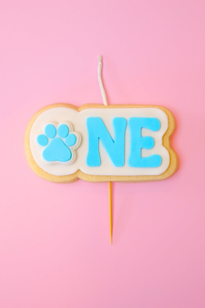 Candle shaped like a cookie with blue 'ONE' and a paw print on a pink background