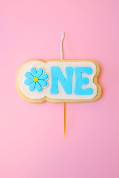 Candle shaped like a cookie with 'ONE' and a flower on a pink background