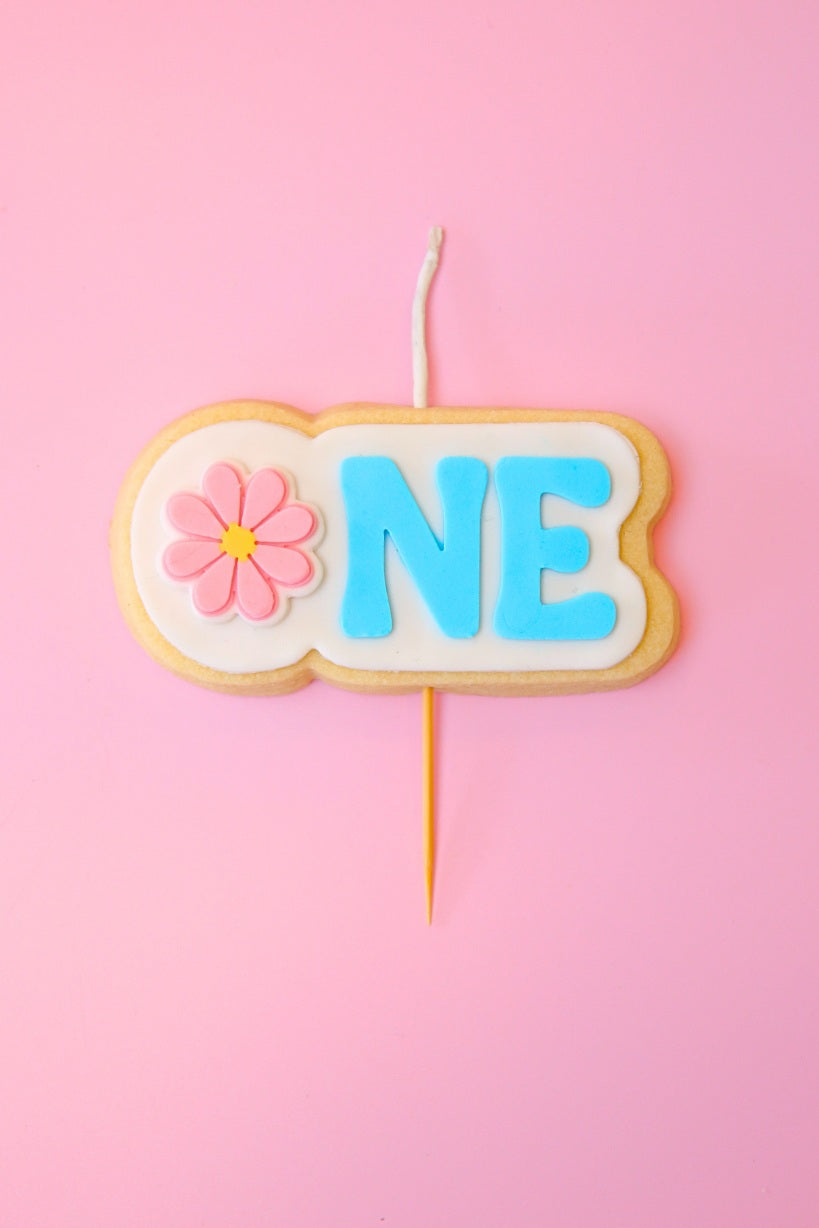 Candle shaped like a cookie with 'ONE' and a flower on a pink background