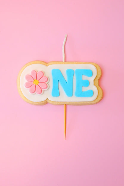 Candle shaped like a cookie with 'ONE' and a flower on a pink background