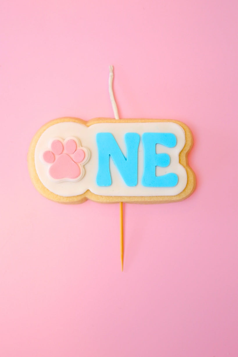 Cookie-shaped candle with pink paw print and blue 'ONE' on a pink background