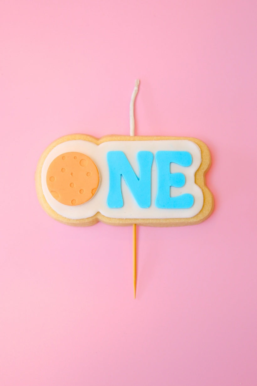 Candle shaped like a cookie with 'ONE' on a pink background with orange planet 