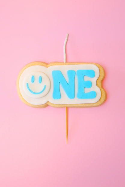 Candle shaped like a cookie with 'ONE' and a blue smiley face on a pink background