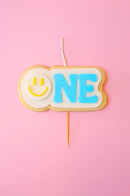 Candle shaped like a cookie with a smiley face and 'ONE' text on a pink background
