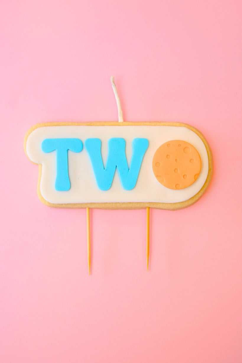 Candle shaped like the number ' TWO ' with blue letters and an orange moon with pink background