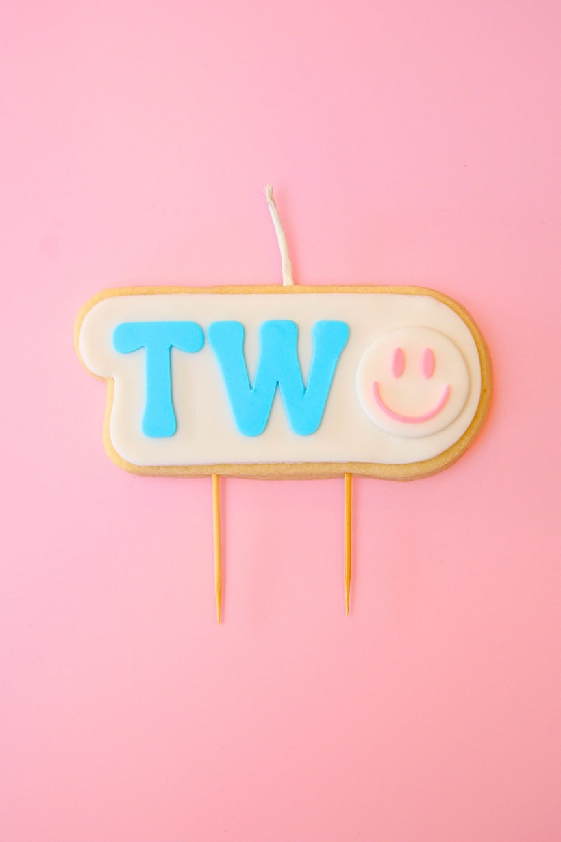 Candle shaped like a cookie with 'TWO' and a smiley face on a pink background