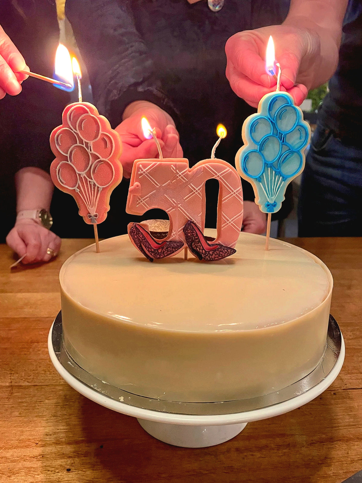 A round beige cake on a white pedestal stand with cookie candles shaped like balloons in pink and blue, and a number 50 candle. Several hands are lighting the candles with matches on a wooden table.