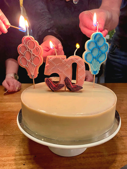 A round beige cake on a white pedestal stand with cookie candle shaped like balloons in pink and blue, and a number 50 candle. Several hands are lighting the candles with matches on a wooden table.