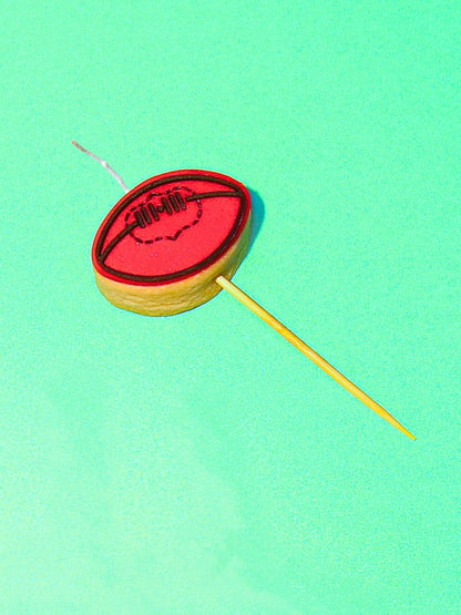 Red fondant football on a sugar cookie with a candle wick. Served on a stick to place in a cake. Photograhped against a green background.