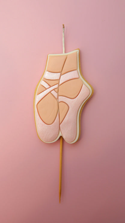 A ballet pointe shoe shaped cookie candle on a wooden stick, set against a solid pink background.