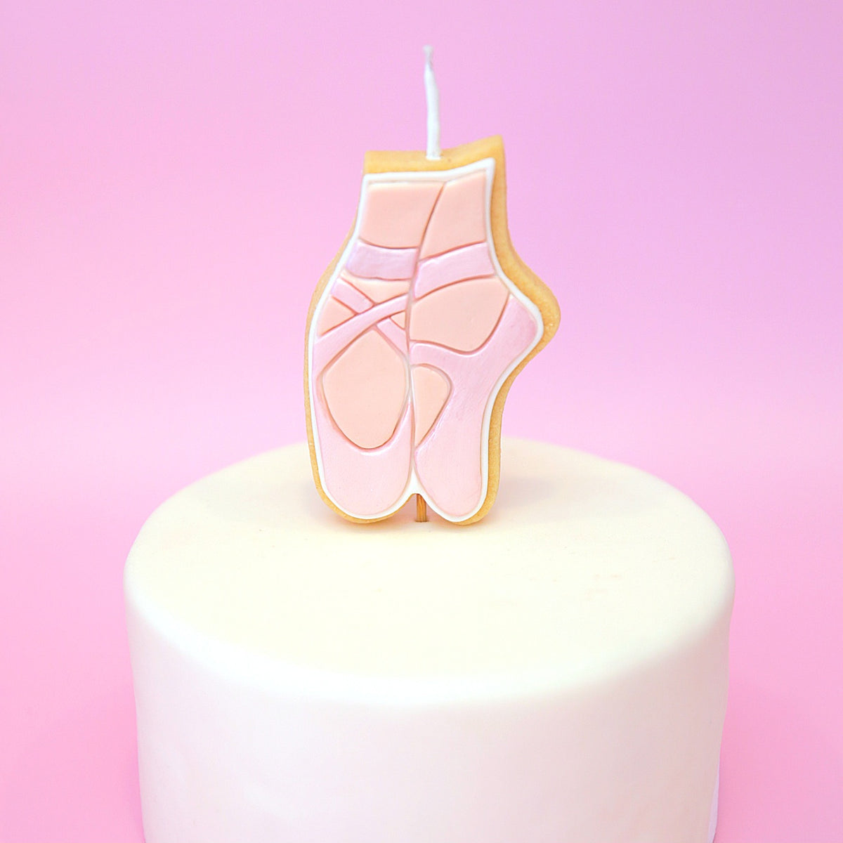 A ballet shoe shaped candle with pink details and a white wick, displayed on top of a white frosted cake against a pink background.