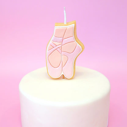 A ballet shoe shaped candle with pink details and a white wick, displayed on top of a white frosted cake against a pink background.