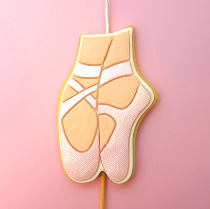A ballet shoe-shaped Cookie Candle decorated with pink icing and white ribbon details, set against a solid pink background.