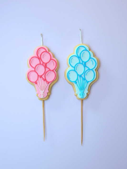 Two balloon-shaped cookie candles, one pink and one blue, on a light purple background.