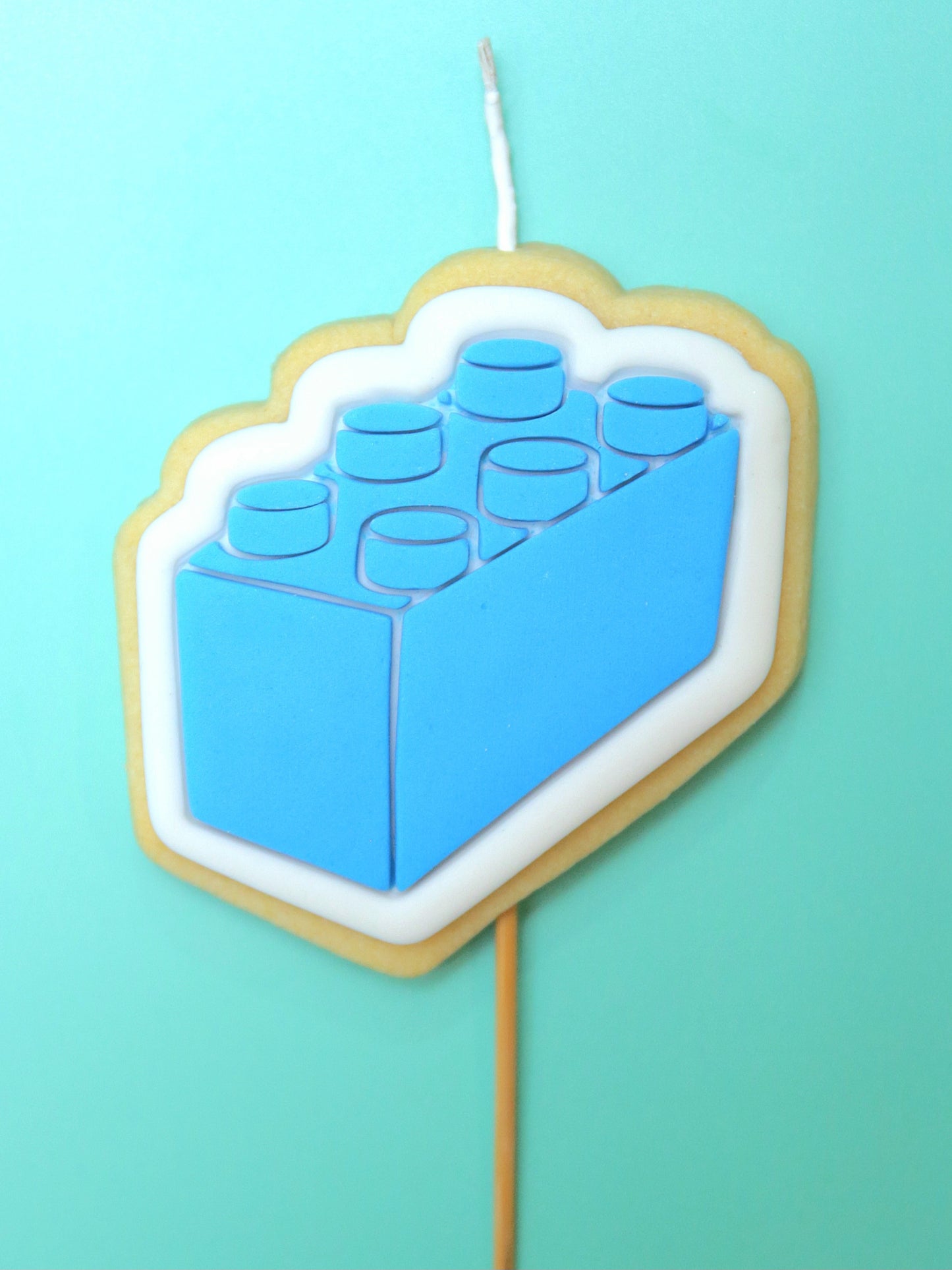 Blue lego block-shaped cookie on a stick with a wick against a teal background