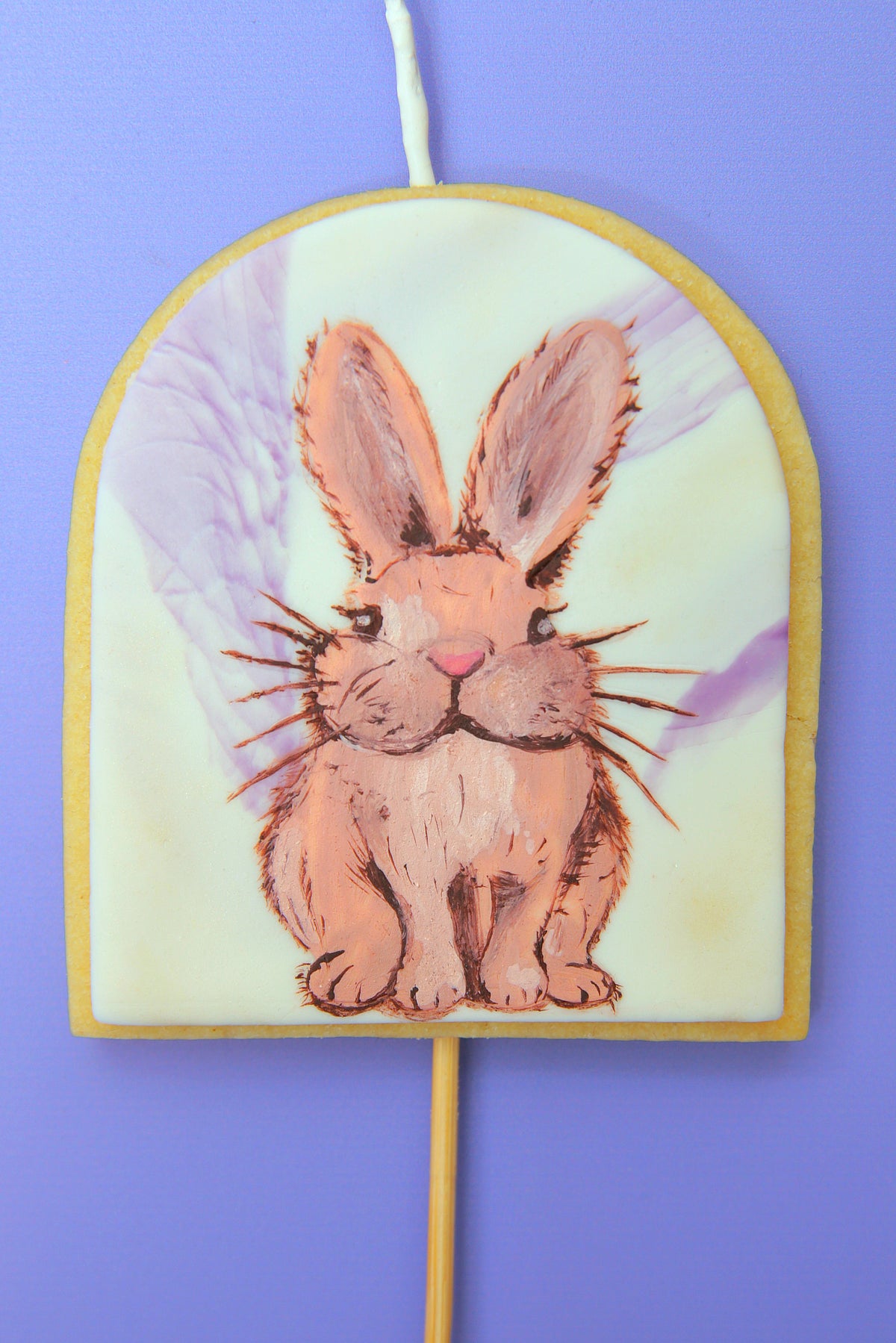 A cookie candle cake topper that is arch shaped with a painted brown bunny on a pale yellow background, mounted on a wooden stick against a solid purple backdrop.