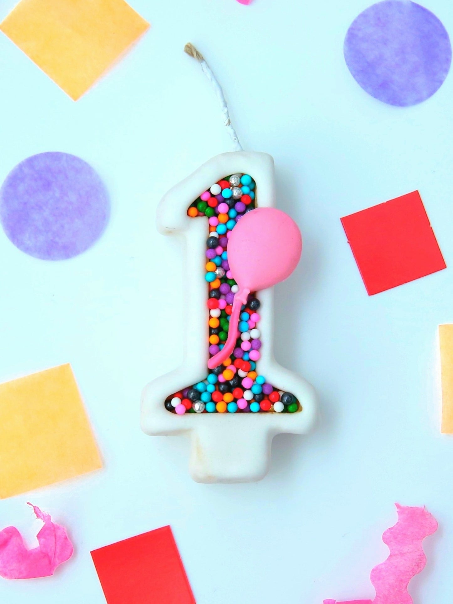 Number 1 birthday candle with colourful sprinkles and a pink balloon on a light blue background with colorful shapes.