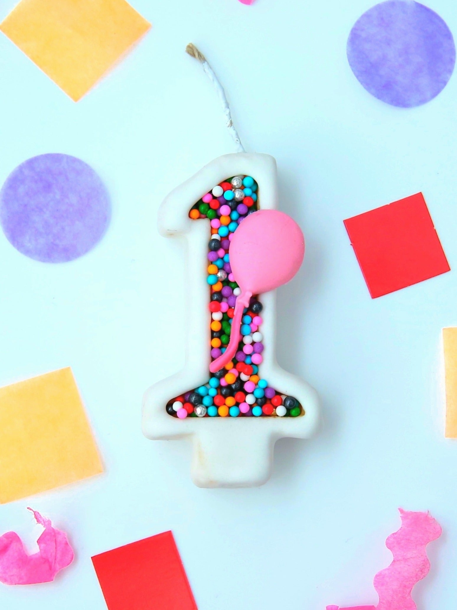 Number 1 birthday candle with colourful sprinkles and a pink balloon on a light blue background with colorful shapes.