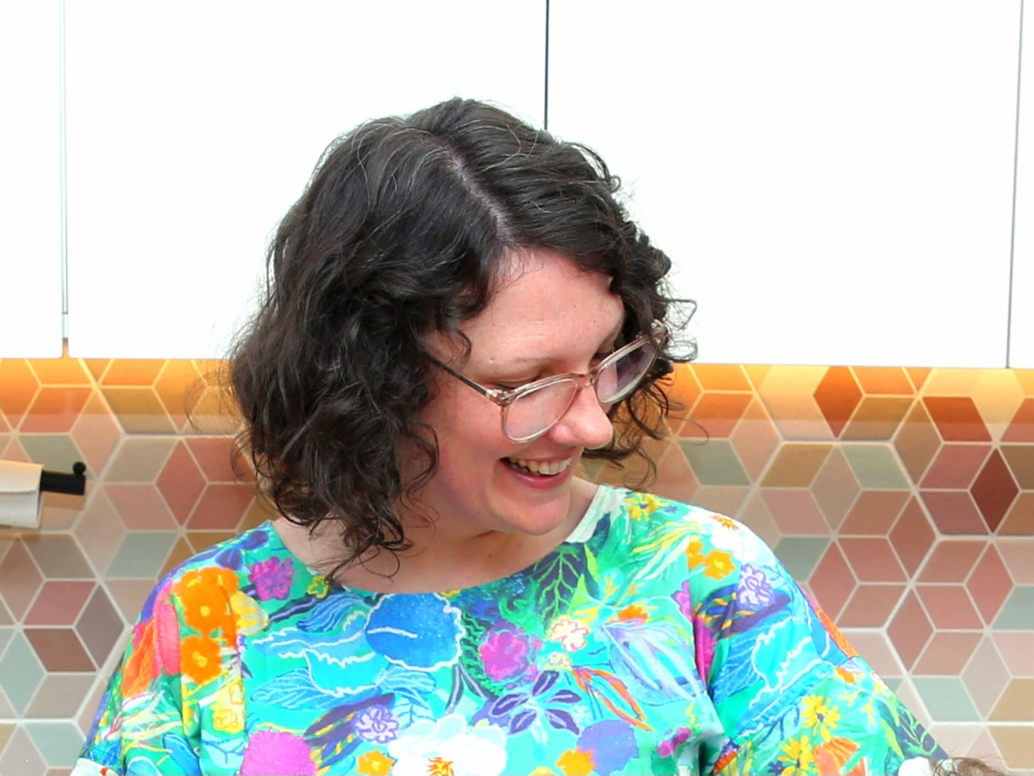 Person smiling wearing a colourful floral dress in a kitchen with a tiled backsplash.