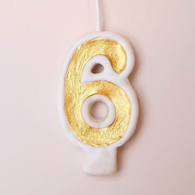 Candle shaped like the number 6 with gold and white design on a light pink background