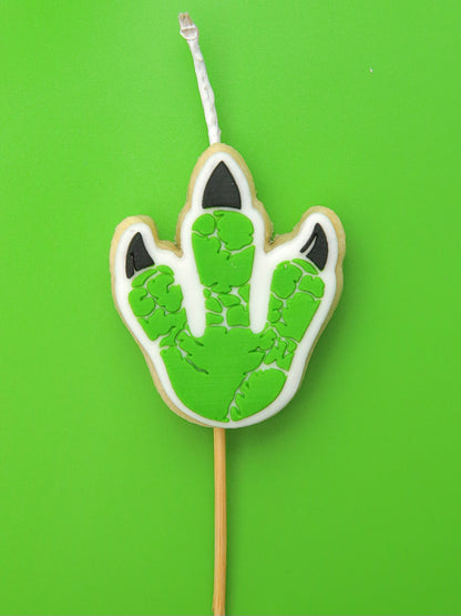 Cookie with green dinosaur footprint design that is also a candle on a stick against a green background