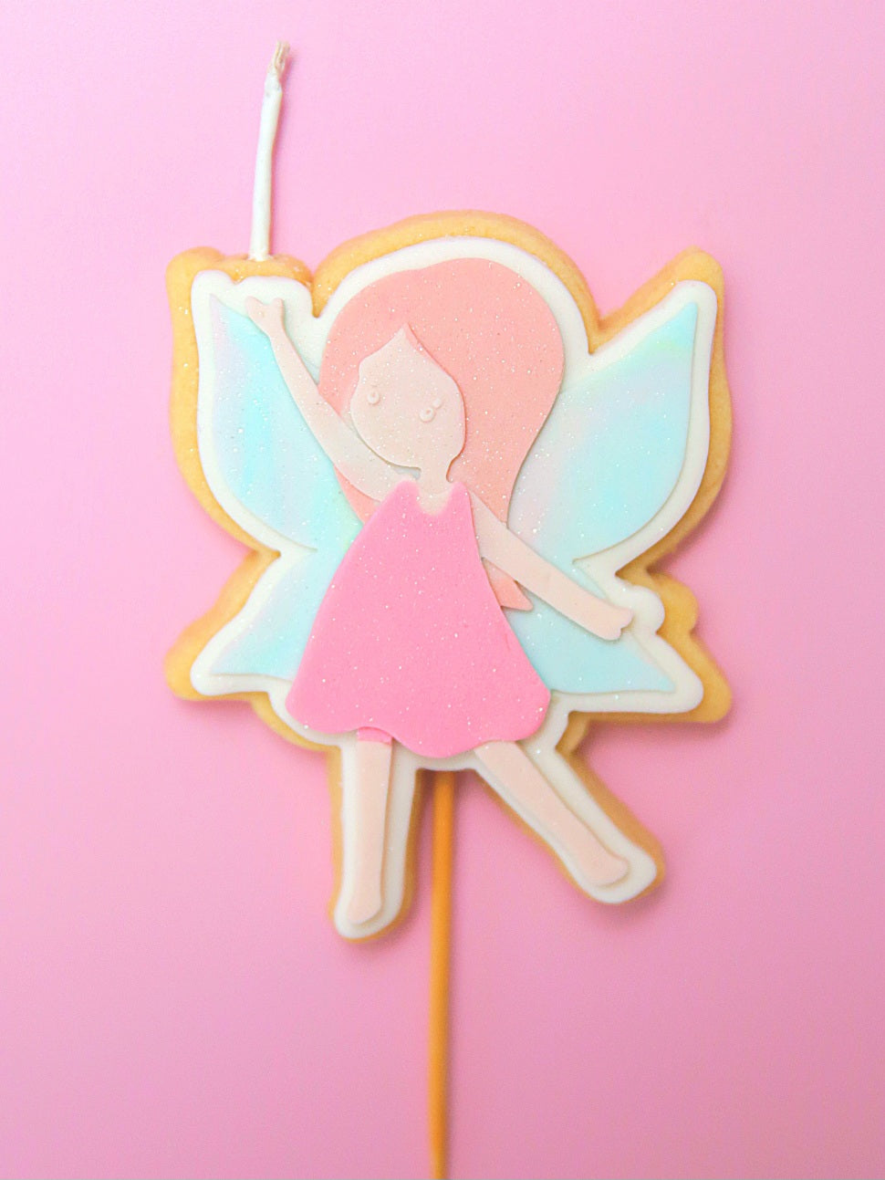 Fairy-themed cookie on a stick and wick with pastel colours and glitter against a pink background