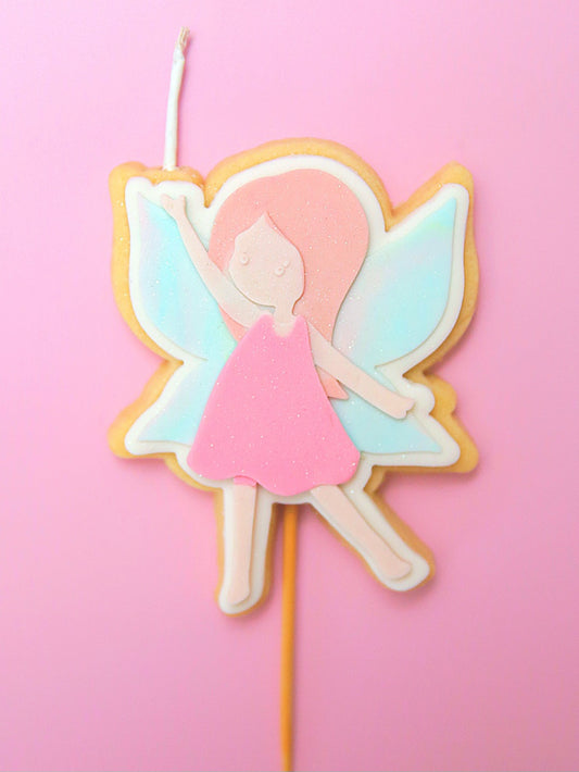 Fairy-themed cookie on a stick and wick with pastel colours and glitter against a pink background