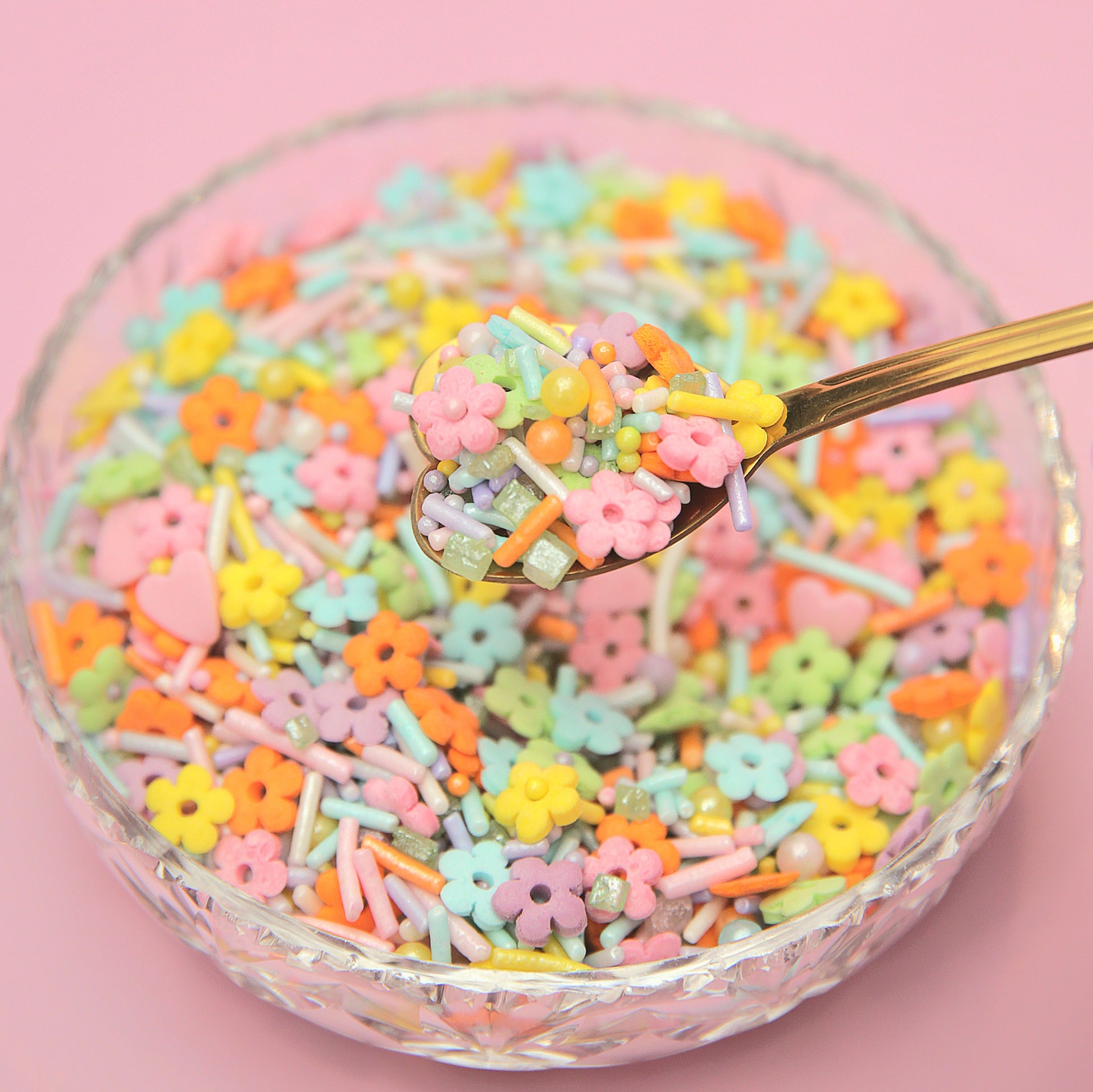 Colorful flower-shaped sprinkles in a glass bowl with a spoon on a pink background