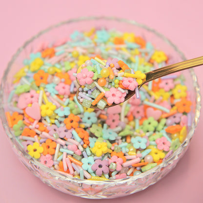 Colorful flower-shaped sprinkles in a glass bowl with a spoon on a pink background