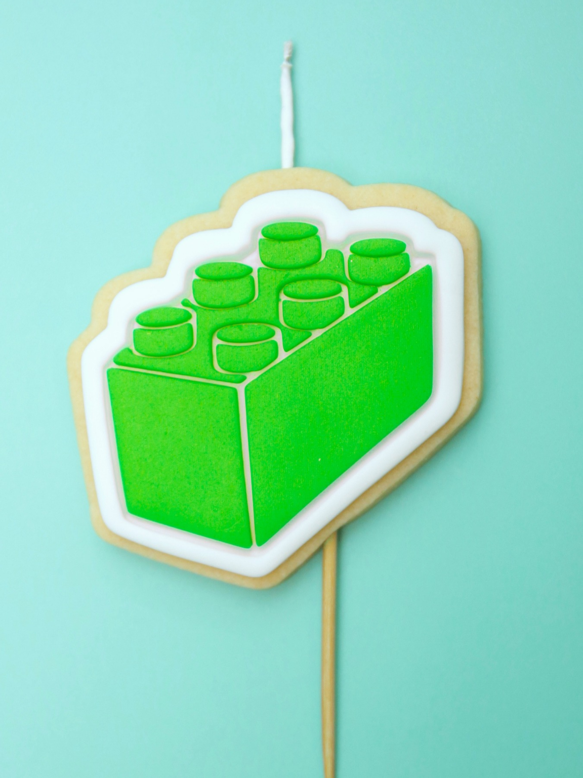 Green block-shaped cookie on a stick with a wick against a light blue background
