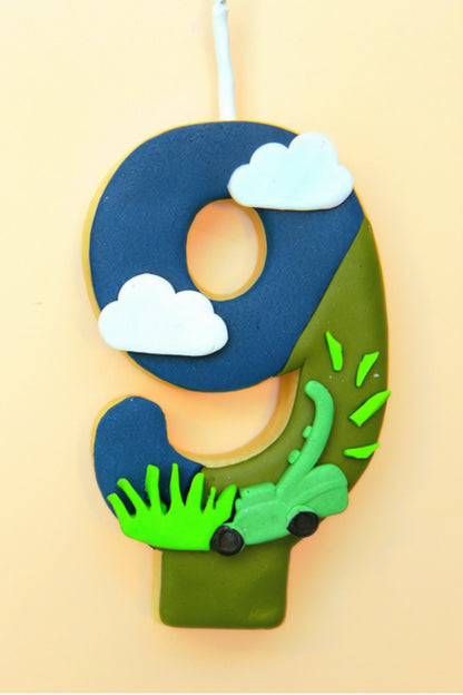 A decorated number 9 birthday cookie or cake topper with a blue and green landscape design, featuring white clouds, bright green grass, and a small green lawn mower, shown against a solid pale peach background.