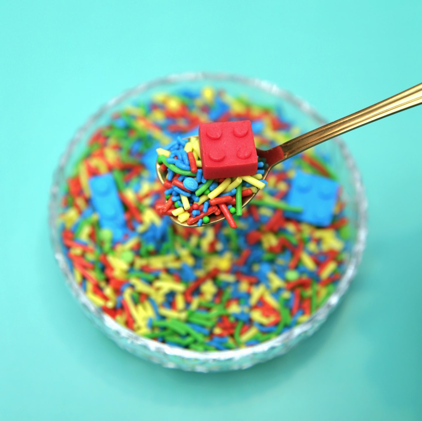 Colorful sprinkles with a red fondant Lego piece on a spoon against a turquoise background