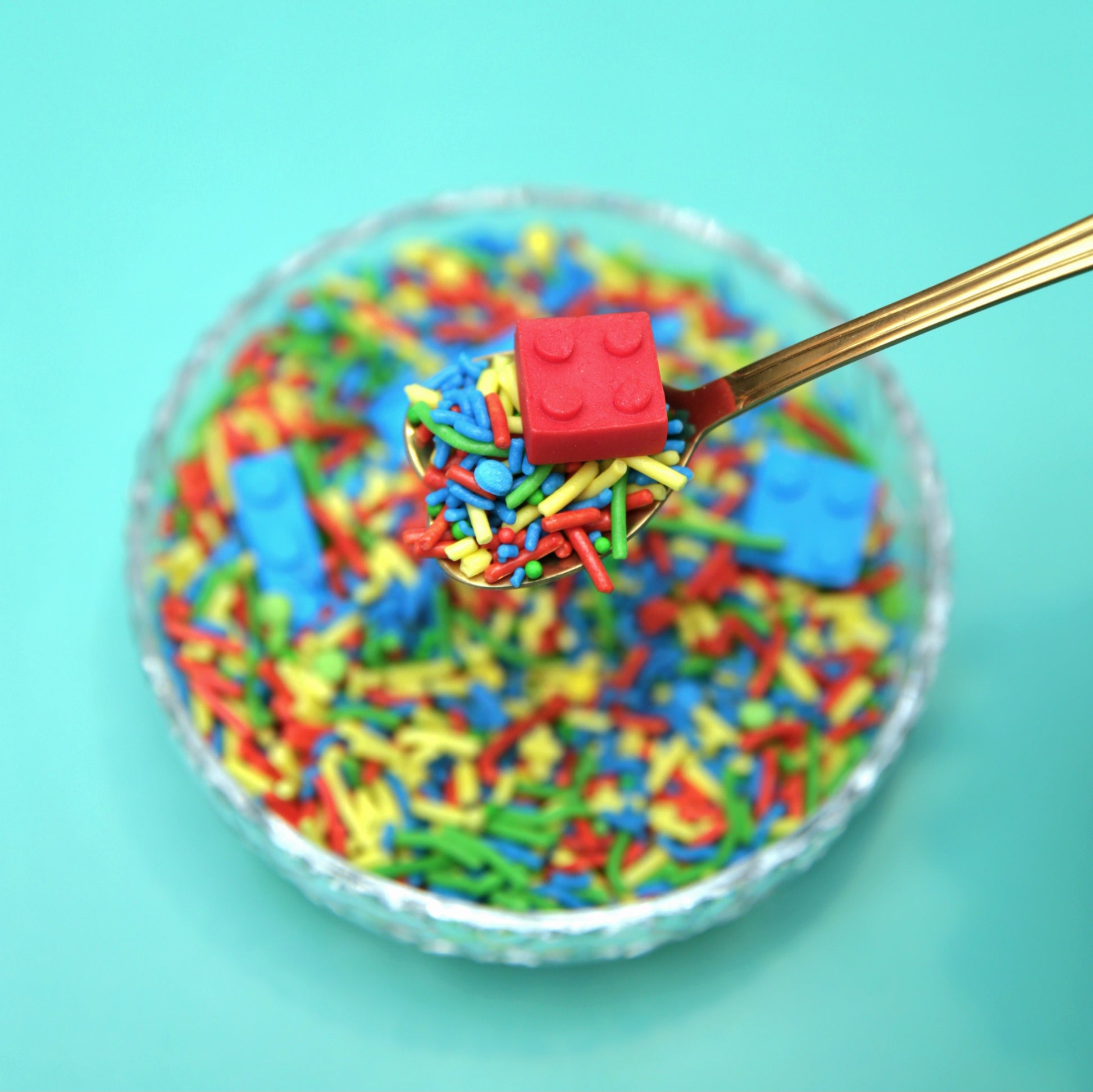 Colorful sprinkles with a red fondant Lego piece on a spoon against a turquoise background