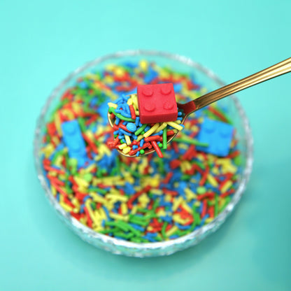 Colorful sprinkles with a red fondant Lego piece on a spoon against a turquoise background