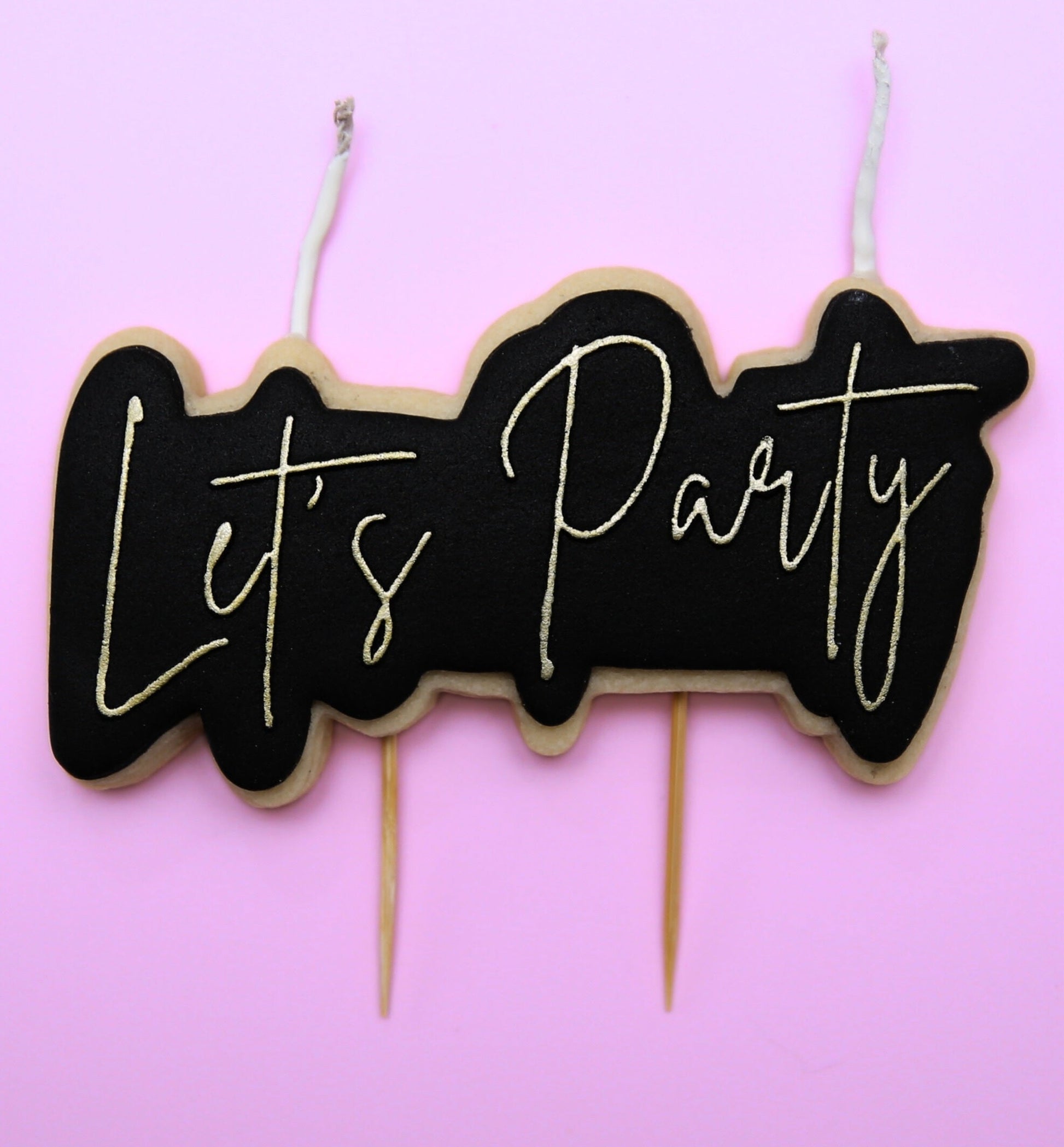 A cookie candle cake topper that reads 'let's party' in gold cursive on a black background. 