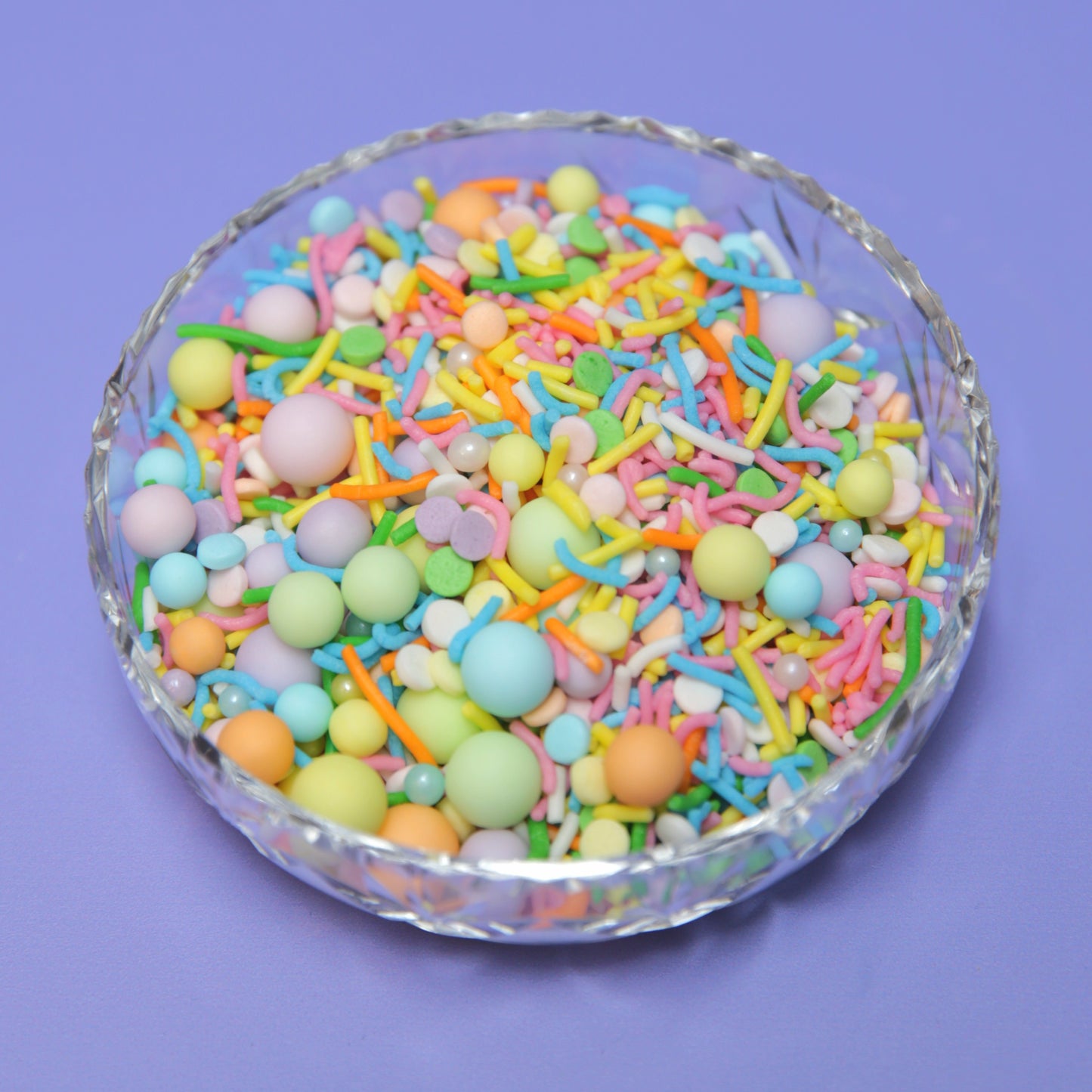 Colorful pastel rainbow sprinkles in a glass bowl on a purple background