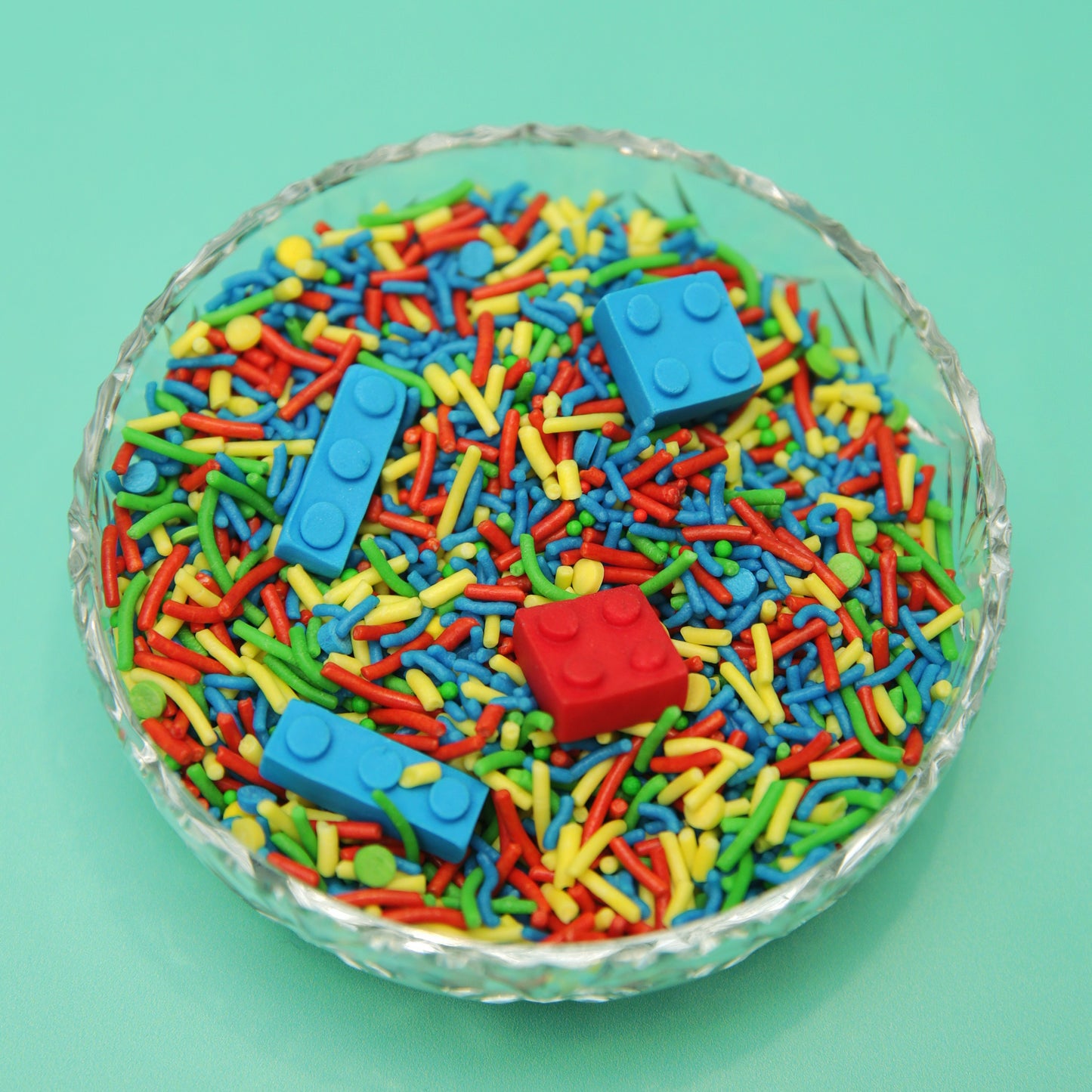 Colorful sprinkles with fondant brick pieces in a glass bowl on a teal background