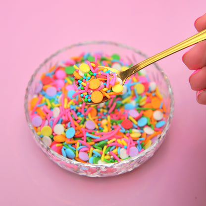 Colorful neon rainbow sprinkles in a glass bowl with a gold spoon on a pink background