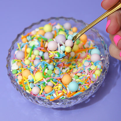 Colorful pastel rainbow sprinkles in a glass bowl with a gold spoon on a purple background