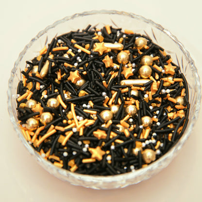 Glass bowl filled with black, gold, and white decorative sprinkles on a beige background