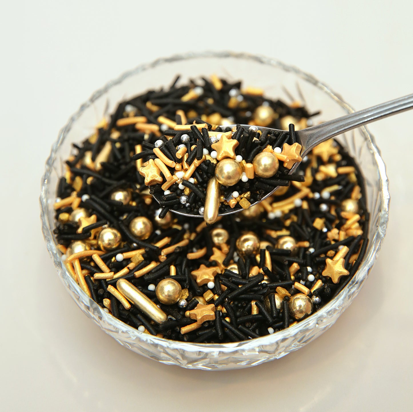 Glass bowl filled with black, gold, and silver star-shaped sprinkles with a spoon.