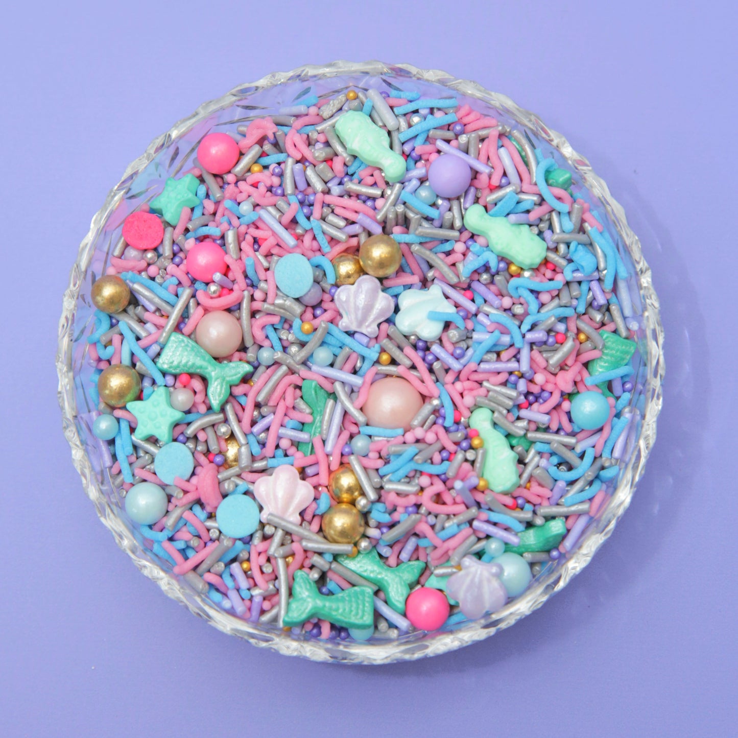 Colorful sprinkles with mermaid-themed decorations in a glass bowl on a purple background