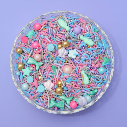 Colorful sprinkles with mermaid-themed decorations in a glass bowl on a purple background