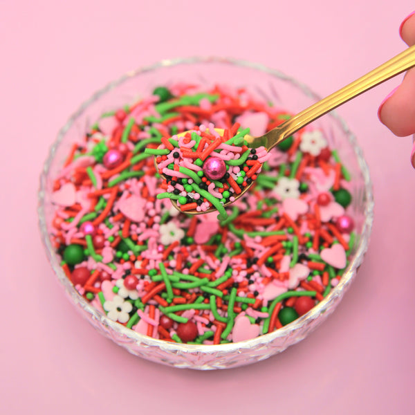 Pink, red and green assorted sprinkles in a glass bowl with a spoon on a pink background