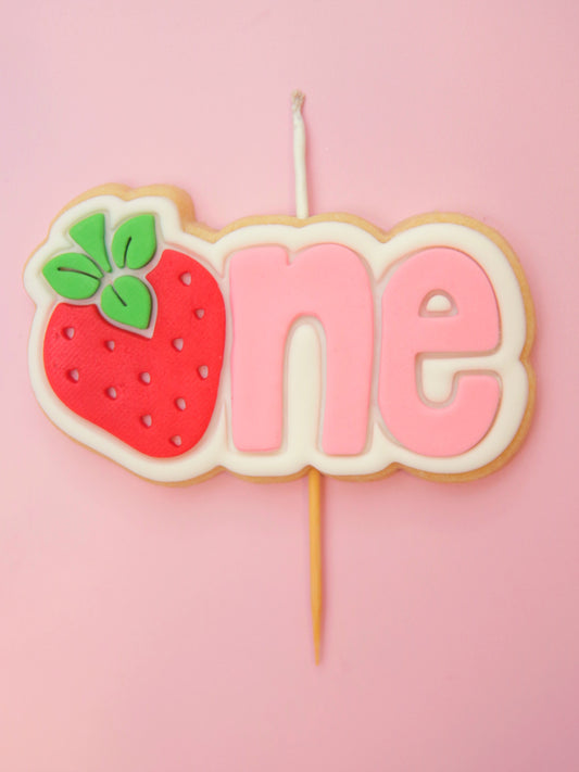 A pink 'one Cookie birthday candle with pink text and a strawberry replacing the 'O'