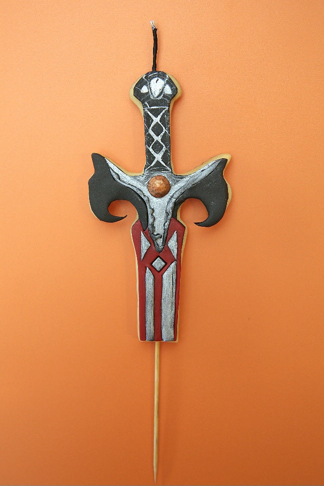 A decorative cake topper shaped like a fantasy sword, featuring a silver and black hilt with red and white detailing on the blade, mounted on a wooden stick against a solid orange background.