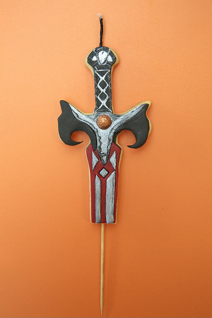 A decorative cake topper shaped like a fantasy sword, featuring a silver and black hilt with red and white detailing on the blade, mounted on a wooden stick against a solid orange background.