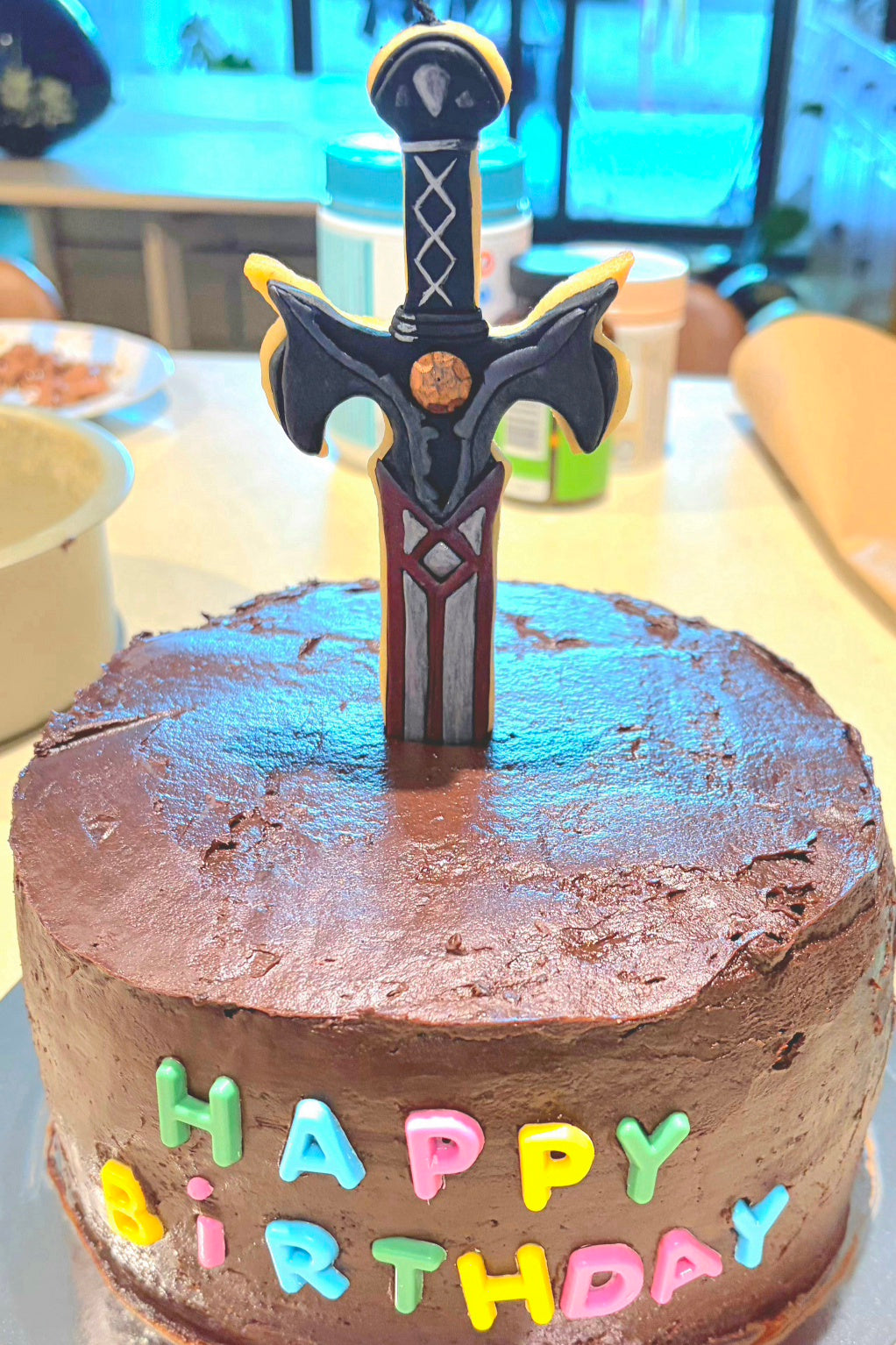 A round chocolate cake with brown frosting, colorful 'HAPPY BIRTHDAY' lettering on the front, and a decorative sword Cookie Candle Cake Topper standing upright on top.