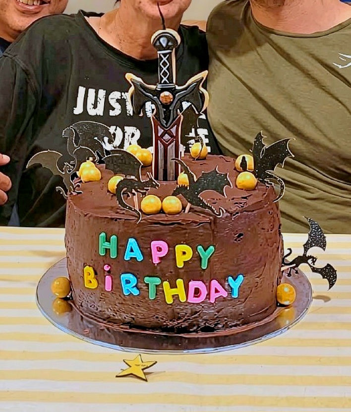 A chocolate birthday cake decorated with colorful 'HAPPY BIRTHDAY' lettering, small orange pom-poms, bat cutouts, and a sword Cookie Candle Cake Topper, shown on a table with two people partially visible behind it.